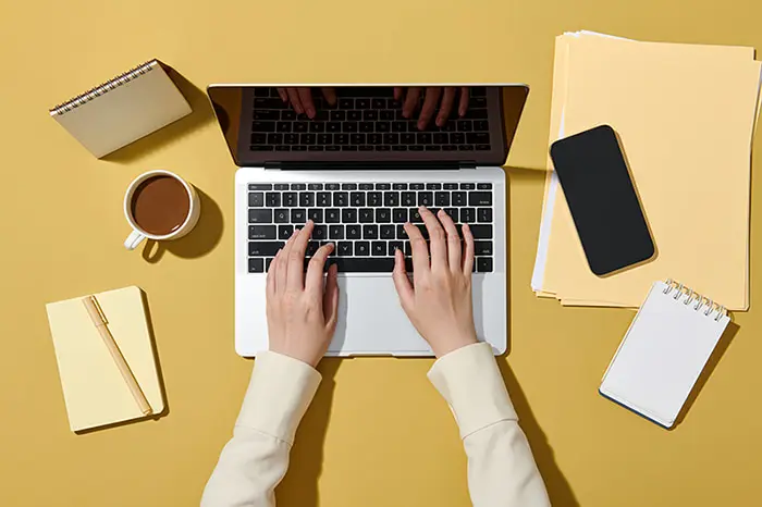 Sur fond marron clair, des mains qui tapent sur un ordinateur portable ouvert. Autour on voit différents éléments comme des feuilles, un bloc notes, une tasse de café, un carnet avec un stylo et un smartphone
