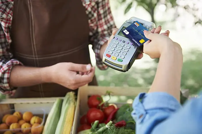 Paiement par carte bancaire sur un marché de fruits et légumes ; on voit le TPE et la partiellement la personne qui le tient ainsi que la carte bancaire dessus et partiellement la personne qui la tient