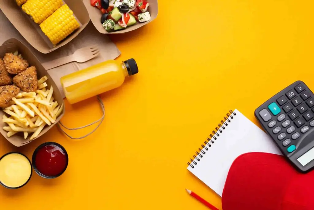 Fond jaune avec des éléments de déjeuner à gauche comme des conteneurs en cartons avec des frites, du mais ou une salade, une fourchette en bois, une bouteille de jus et des sauces. Sur le coin inférieur droit un bloc notes, une calculatrice, une casquette rouge et un crayon.