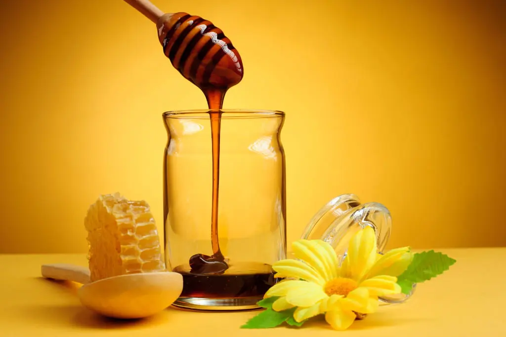 Pot de miel avec miel qui coule, posés sur une table avec une fleur et une cuillère et un morceau de miel, le tout principalement en jaune