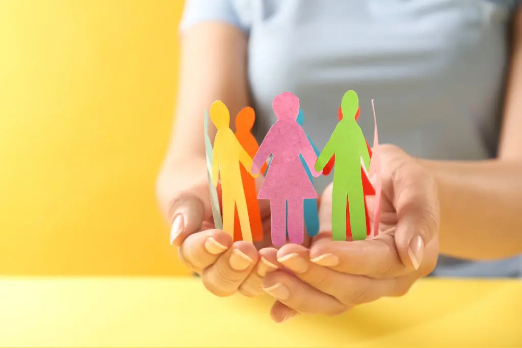 Personnes en papier se tenant la main en cercle dans les mains d'une personne dont on voit seulement le buste et les mains au dessus d'une table, le tout sur fond jaune et table jaune