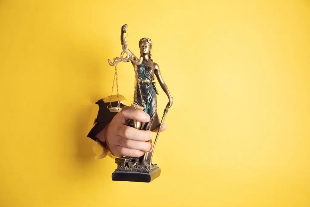 Une main tenant une statuette de l'allégorie de la justice passe à travers ce qui semble être un fond jaune en papier.