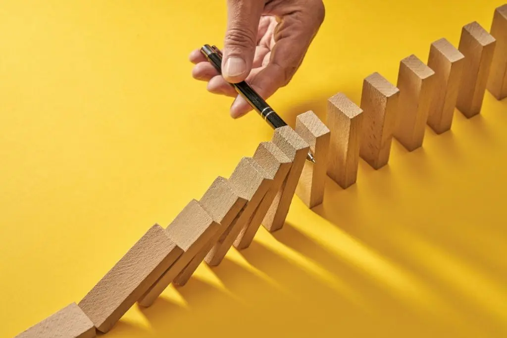 Sur fond jaune, une main glisse un stylo dans une ligne de dominos pour empêcher les derniers de tomber
