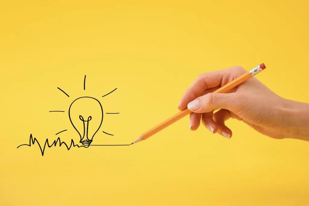 Sur fond jaune, une main tiens un crayon à droite de l'image, qui dessine une ampoule à gauche de l'image