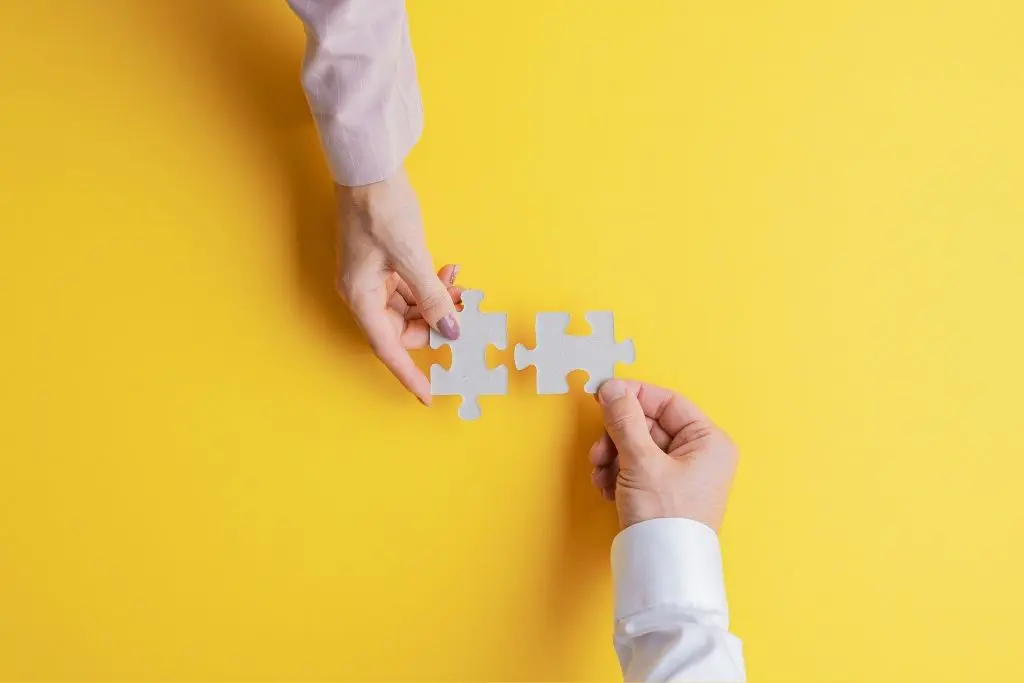 Sur fond jaune, deux mains se tendent pour assembler des pièces de puzzle blanches au centre de la photo