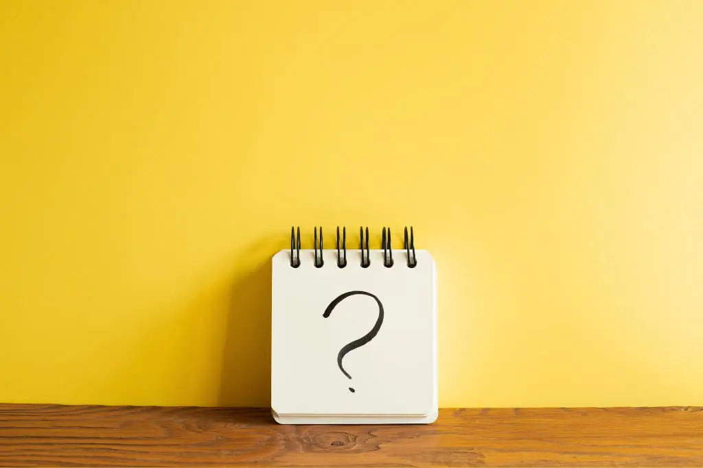 Sur fond jaune et posé sur une table en bois, un bloc notes carré blanc avec un point d'interrogation dessus