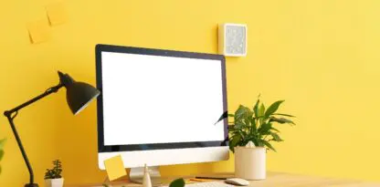 Sur fond jaune, une photo d'un bureau avec un moniteur au fond blanc, une lampe, divers objects de bureau posés et des plantes