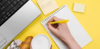 Sur un bureau fond jaune, un coin d'ordinateur portable, une viennoise, une boisson chaude tenue par une main, une autre main qui écrit sur un bloc-notes et un post-it avec écrit "To-do : Choisir les meilleurs titres-restaurant"