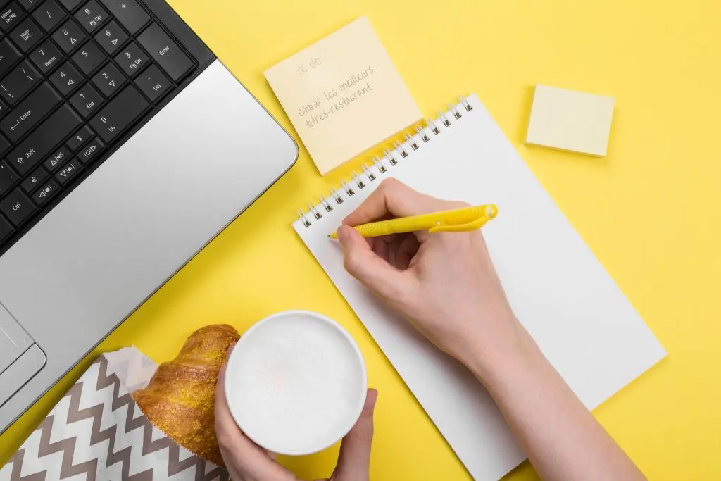 Sur un bureau fond jaune, un coin d'ordinateur portable, une viennoise, une boisson chaude tenue par une main, une autre main qui écrit sur un bloc-notes et un post-it avec écrit "To-do : Choisir les meilleurs titres-restaurant"