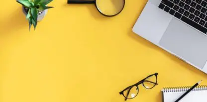 Sur fond jaune vue du dessus d'un ordinateur portable, une plante, une loupe, un carnet et des lunettes de vue.