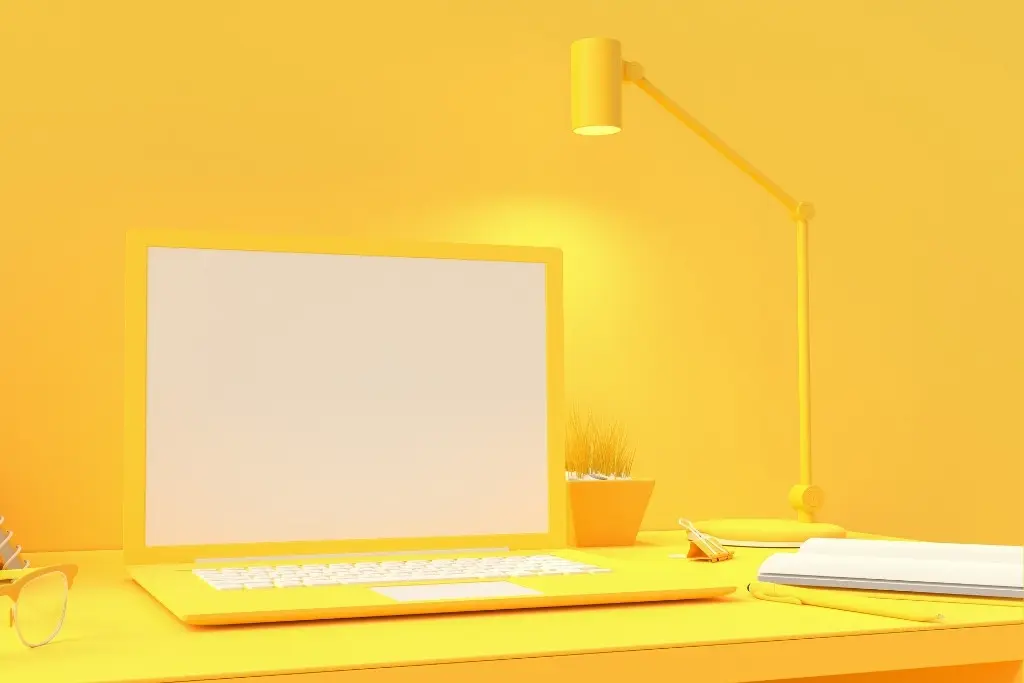 Photo monochromatique jaune d'un bureau avec un ordinateur portable, une lampe, une plante, un carnet et des lunettes de vue.