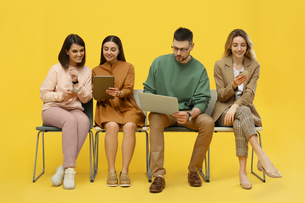 Sur fond jaune, des jeunes femmes et hommes sont assis sur des chaises et discutent en regardant leurs ordinateurs ou smartphones, en attendant leur entretien de recrutement