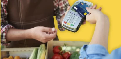 Photo collée sur un fond jaune, d'un paiement par carta bancaire au marché (devant des caisses de fruits et légumes)
