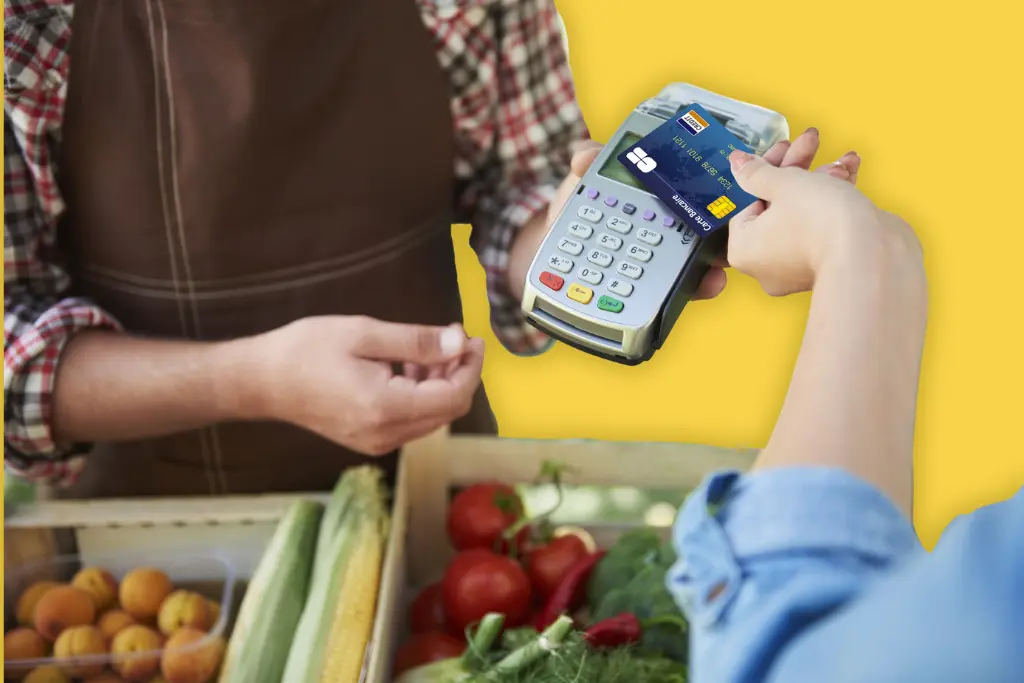 Photo collée sur un fond jaune, d'un paiement par carta bancaire au marché (devant des caisses de fruits et légumes)
