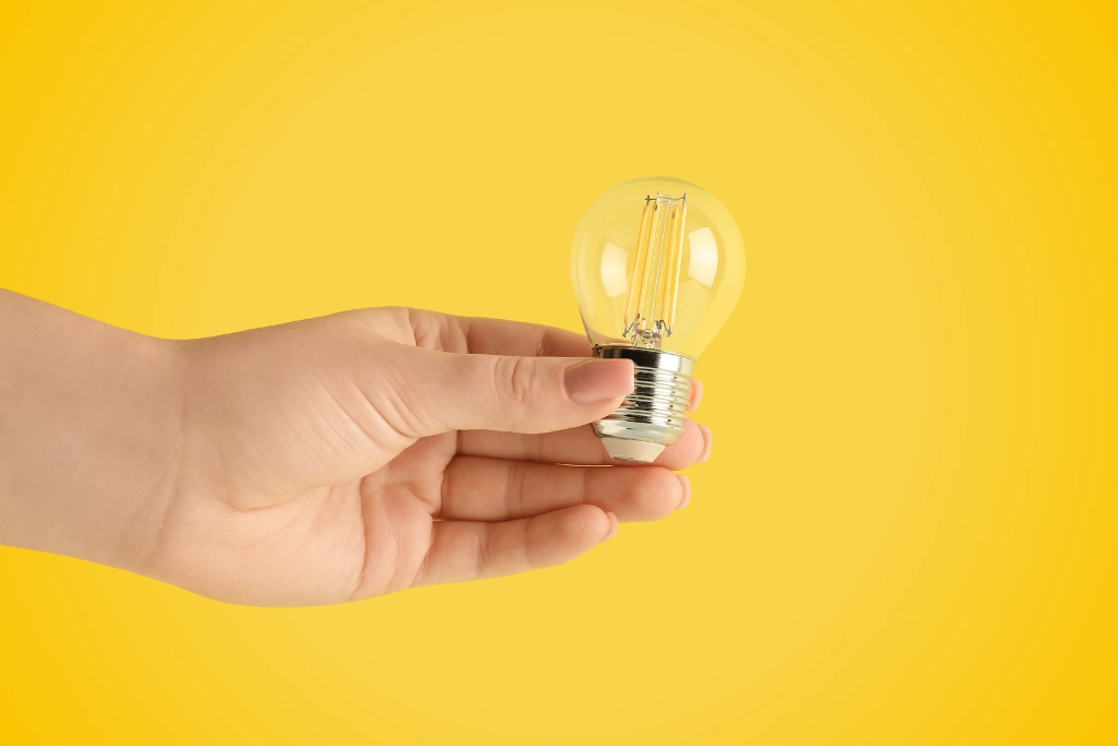 Sur fond jaune, photographie en gros plan d'une main tenant une petite ampoule
