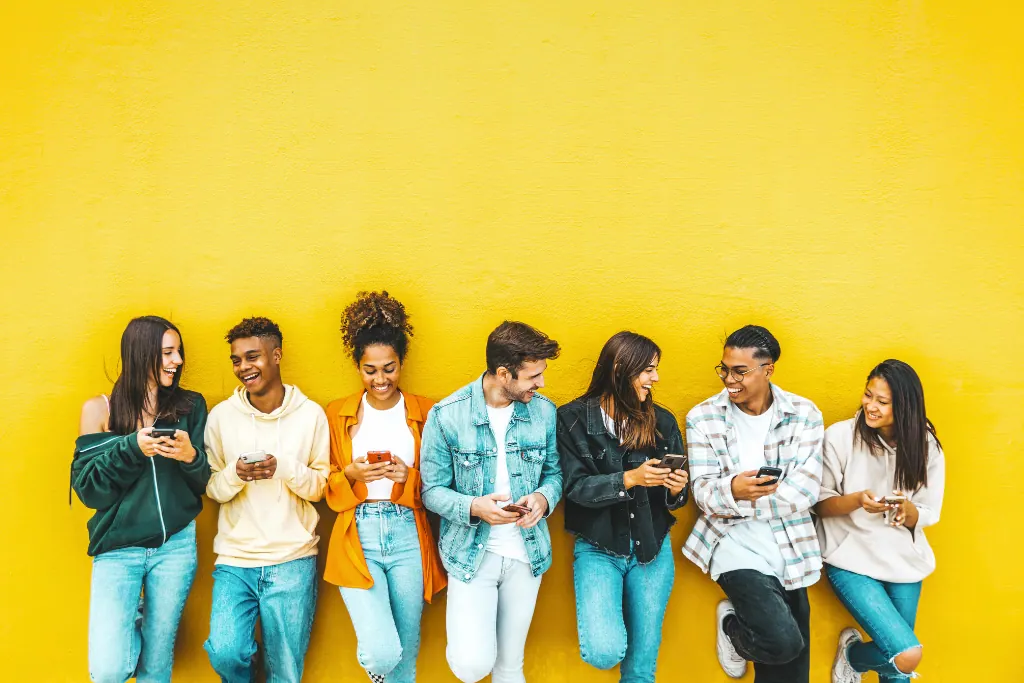 Photographie d'un groupe de jeunes gens souriants adossés à un mur jaune, sur leurs téléphones et échangeant des sourires entre eux