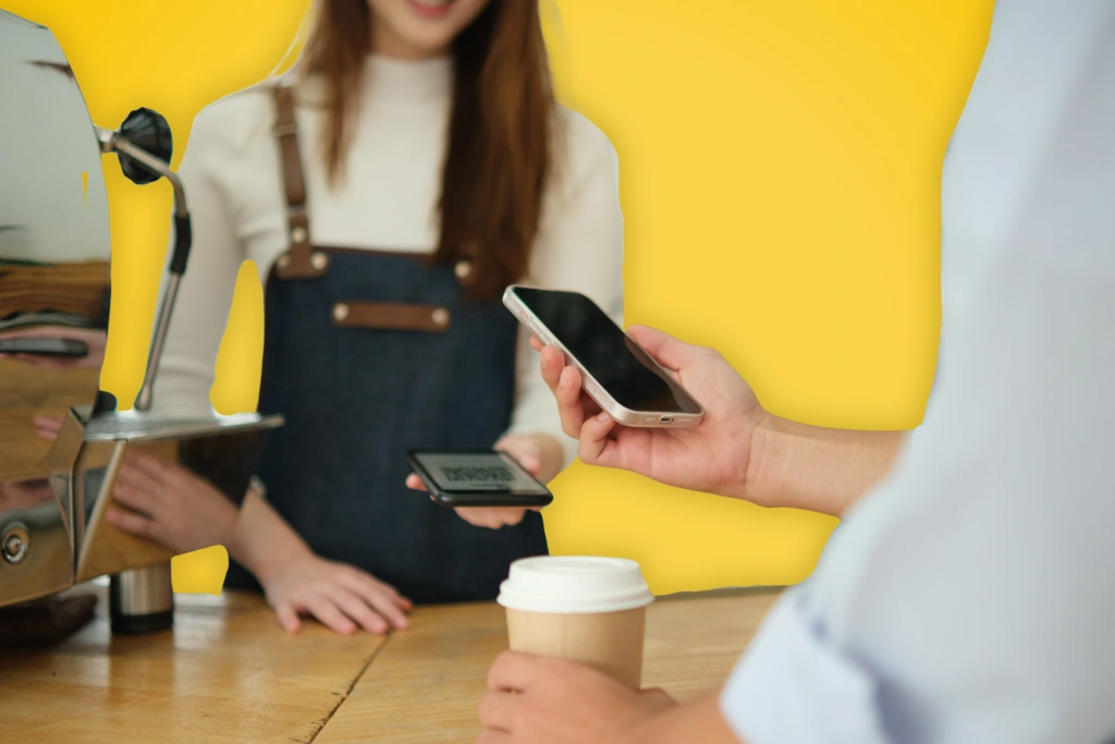 Sur fond jaune, une photo d'une main tendue tenant un smartphone pour payer, face à une employée de café
