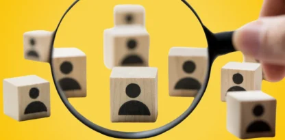Sur fond jaune, une main tient une loupe sur des blocs en bois, sur lesquels il y a des silhouettes de bustes dessinés.