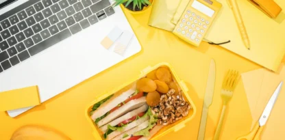 Sur fond jaune sont posés un ordinateur portable, une boîte déjeuner, des bananes et plusieurs éléments de bureau comme une calculatrice ou des feuilles et ciseaux.