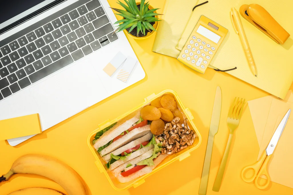 Sur fond jaune sont posés un ordinateur portable, une boîte déjeuner, des bananes et plusieurs éléments de bureau comme une calculatrice ou des feuilles et ciseaux.