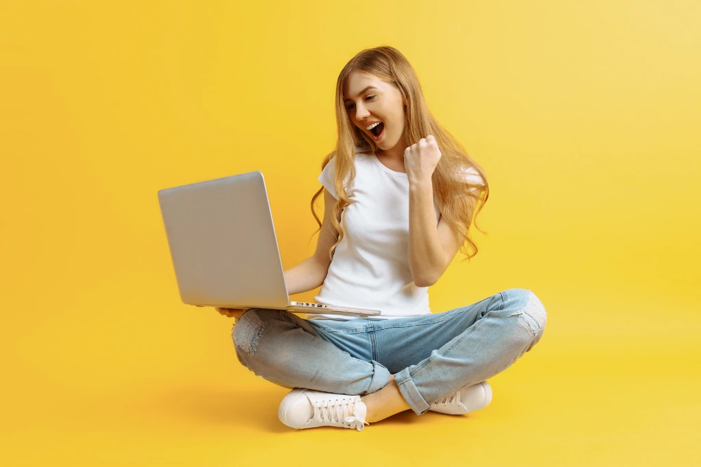 Sur fond jaune, une jeune femme assise en tailleur s'exclame de joie devant son ordinateur portable