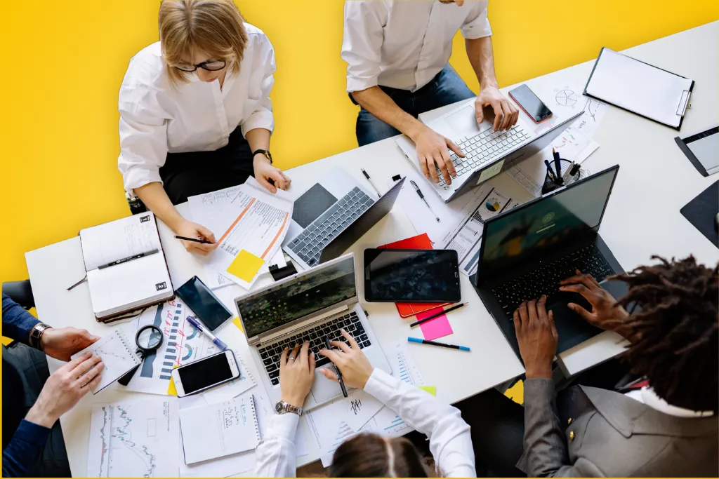 Sur fond jaune, une photographie vue du dessus d'une équipe qui travaille ensemble à une grande table