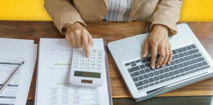 Sur fond jaune, une photographie vue du dessus d'une personne travaillant à son bureau avec une calctulatrice, un ordinateur portable et des documents papier