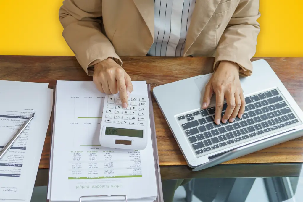 Sur fond jaune, une photographie vue du dessus d'une personne travaillant à son bureau avec une calctulatrice, un ordinateur portable et des documents papier
