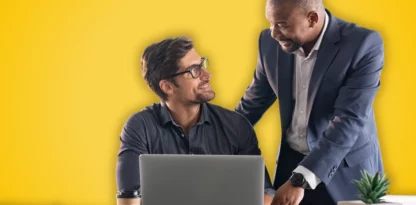 Sur fond jaune, deux hommes se sourient, l'un est assis à son bureau et l'autre est debout à sa droite et pose sa main sur son épaule.