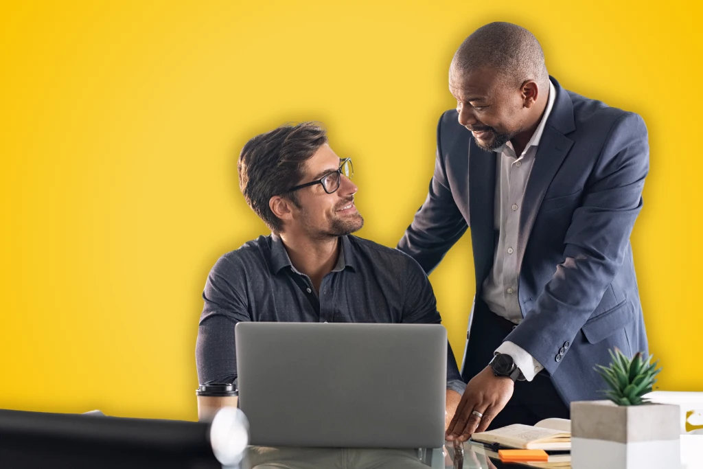 Sur fond jaune, deux hommes se sourient, l'un est assis à son bureau et l'autre est debout à sa droite et pose sa main sur son épaule.